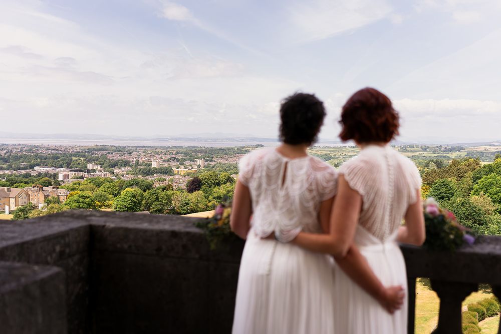 Two brides in white dresses embrace on a stone terrace overlooking a lush green cityscape on their wedding day.