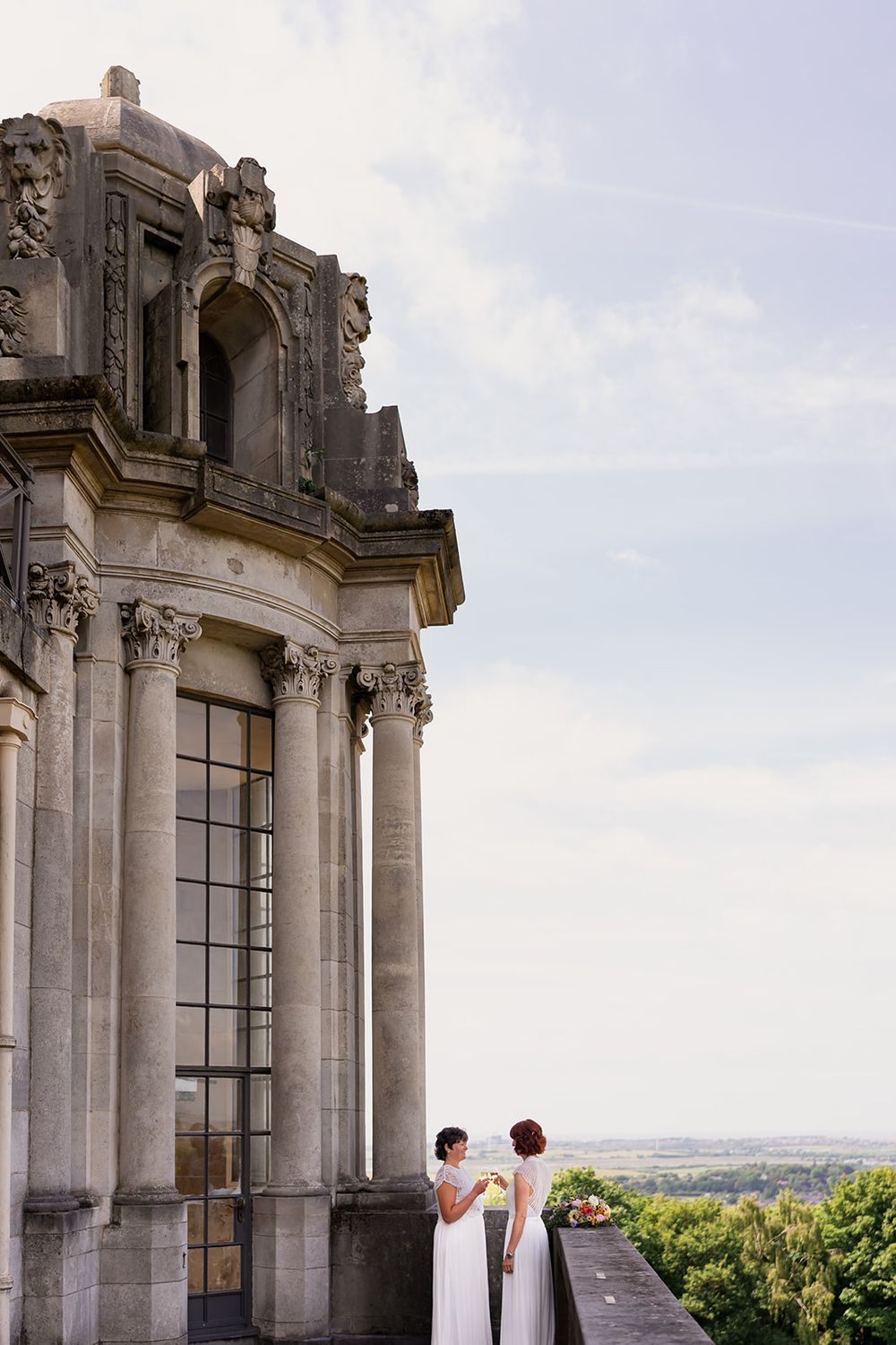 Two brides in white dresses stand on a balcony of an ornate historic stone building overlooking a scenic landscape.