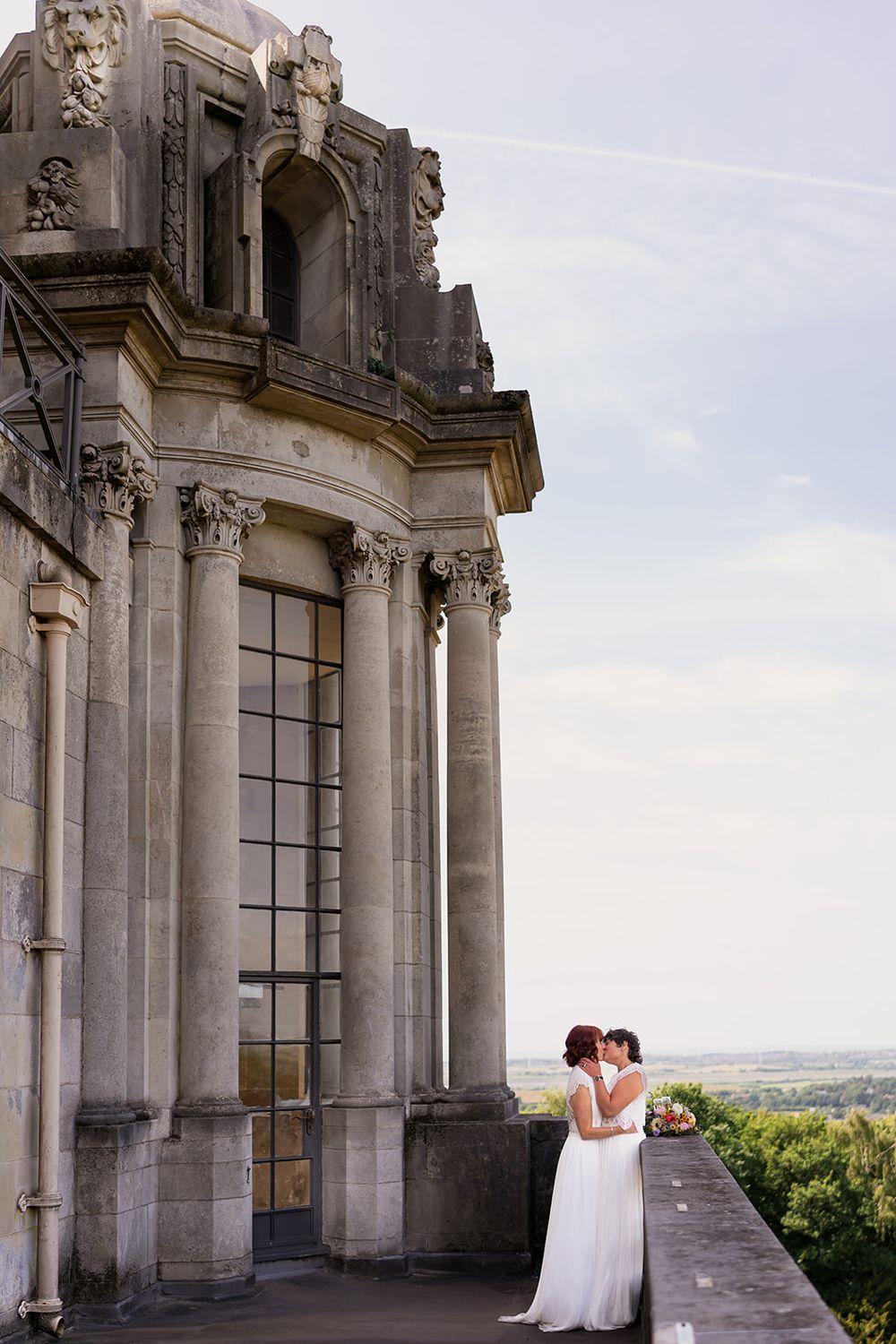 Two brides share a romantic moment beside an ornate stone building on a scenic outdoor terrace.