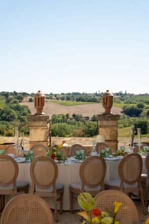 Elegant outdoor dining table with cane chairs set against a scenic Tuscan countryside landscape under a clear blue sky.