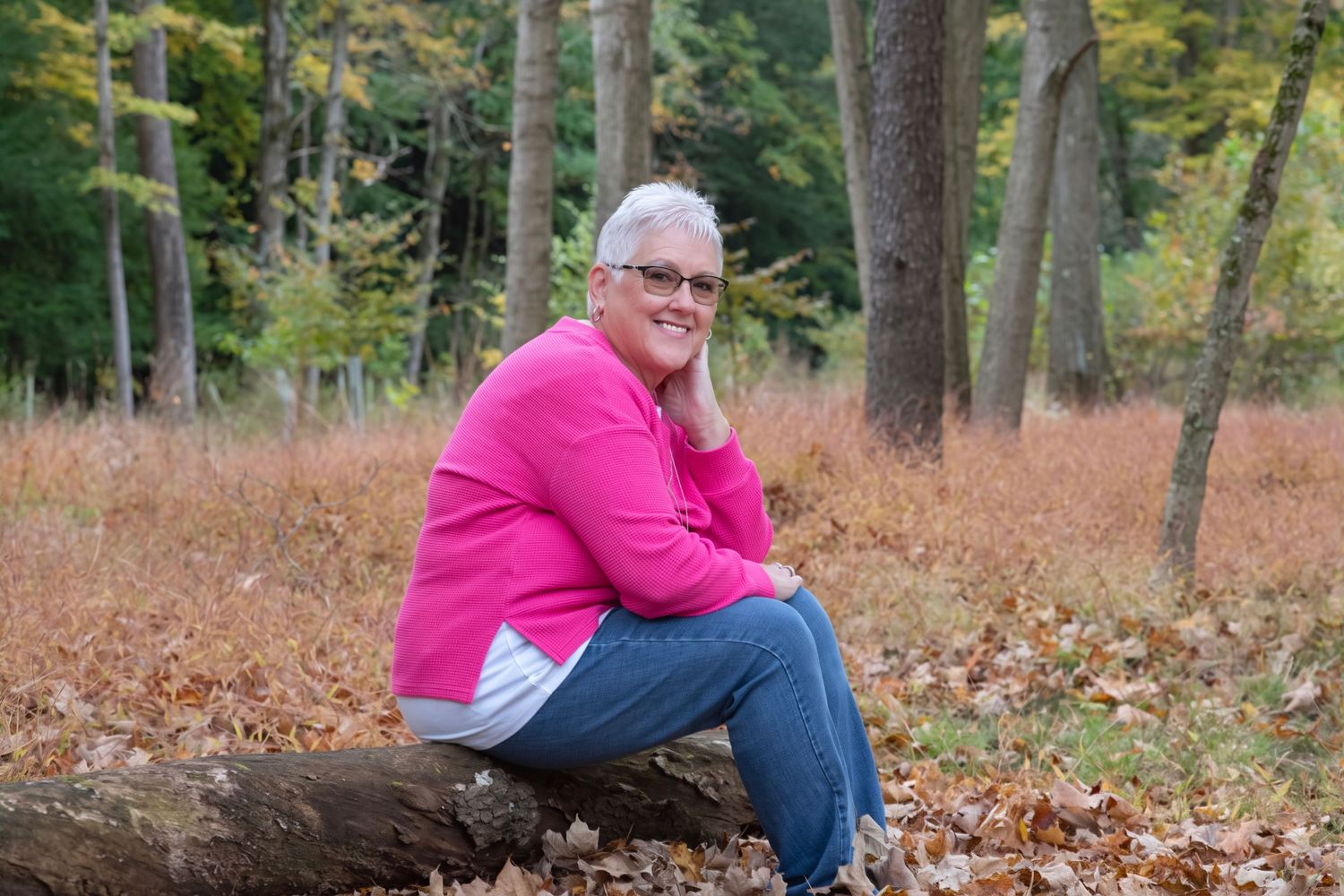 Woman in pink sweater sitting on a log in an autumn forest with colorful foliage and tall trees.