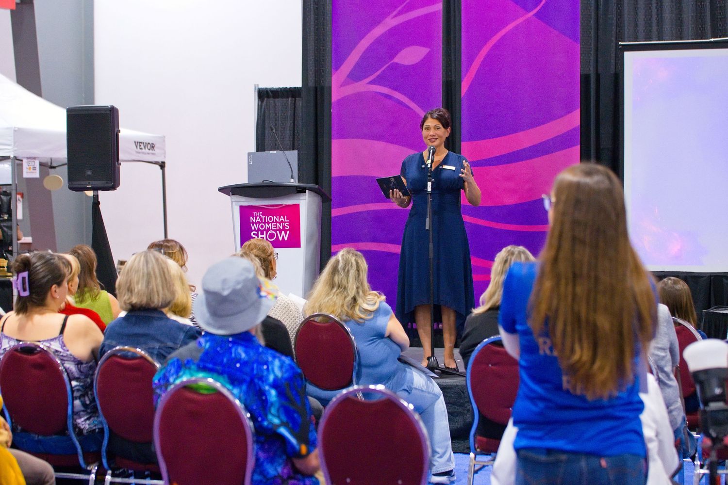 Woman in a blue dress speaks into a microphone on stage to a seated audience at a conference, with purple banners behind her.