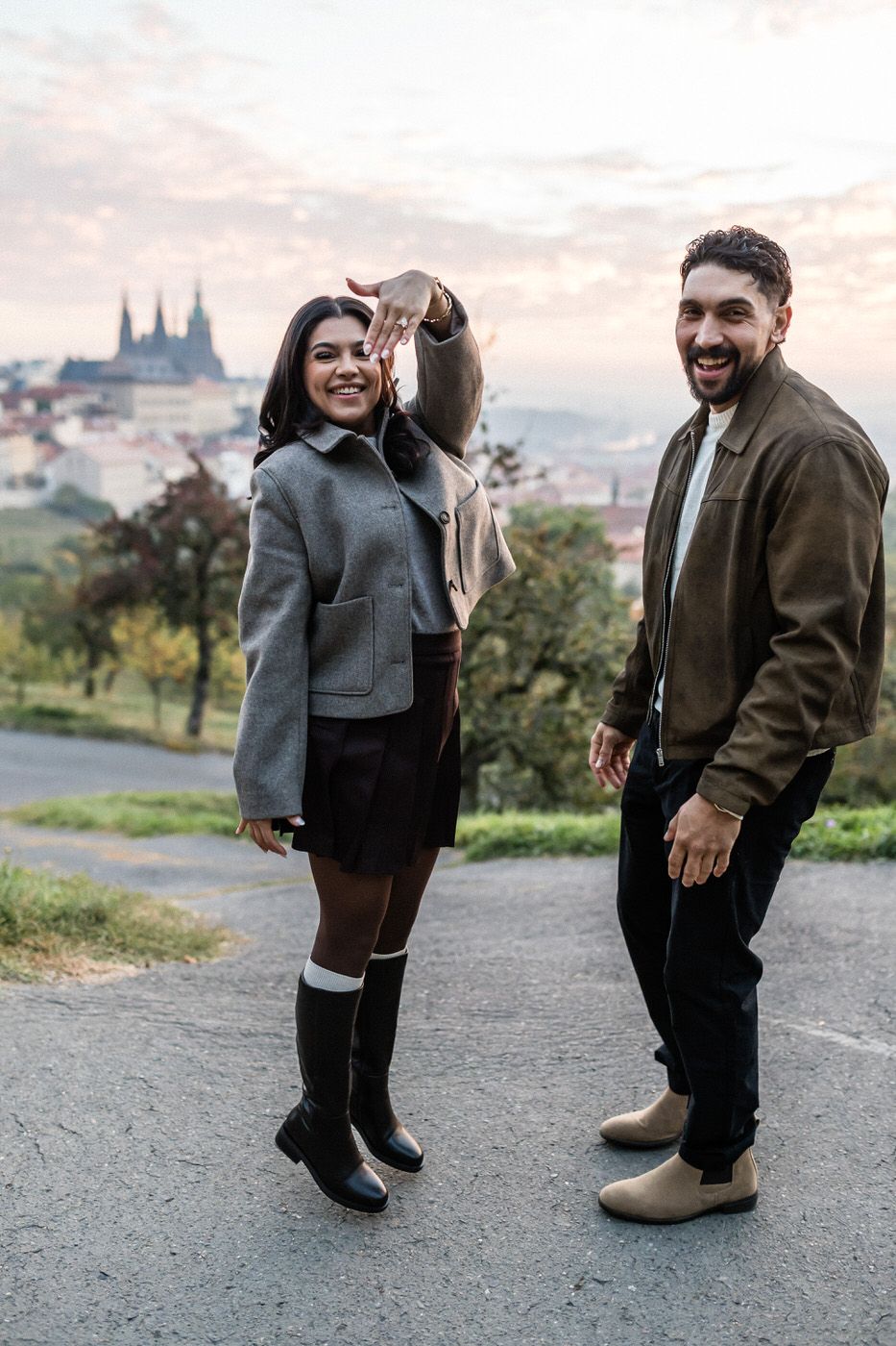 Couple embracing on a hilltop terrace at sunrise with a pastel sky and red-roofed city and castle spires in the background.