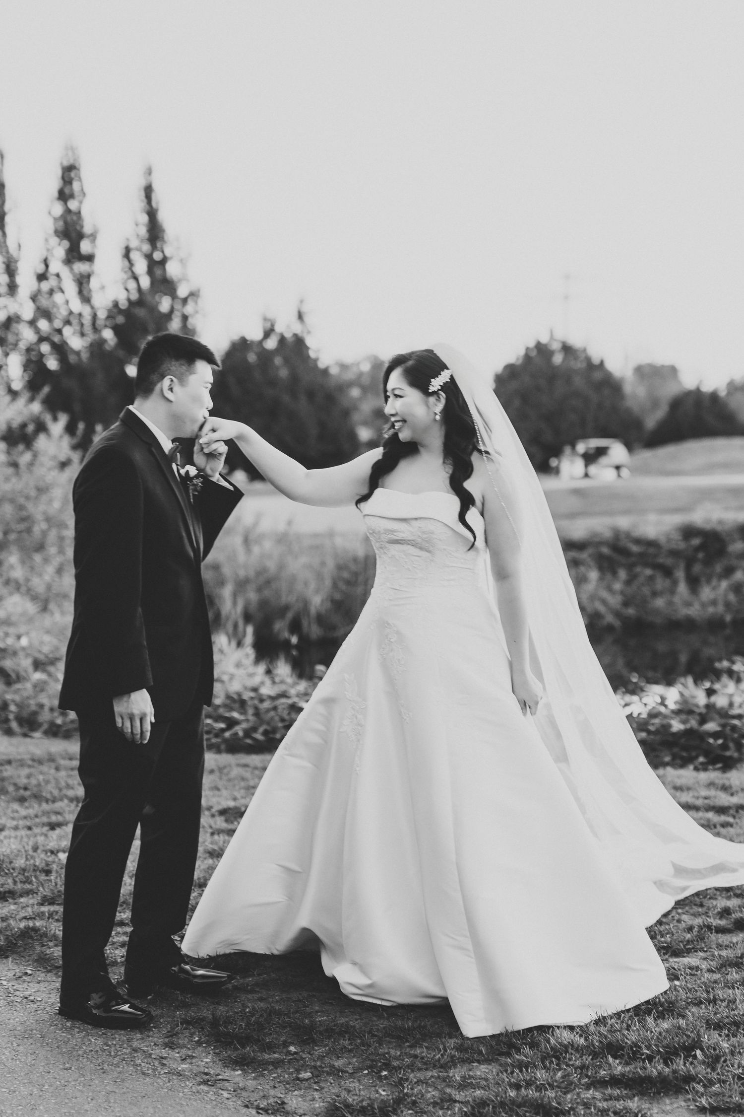 Black and white wedding photo of a groom in a suit kissing the hand of a smiling bride in a strapless gown and veil outdoors.