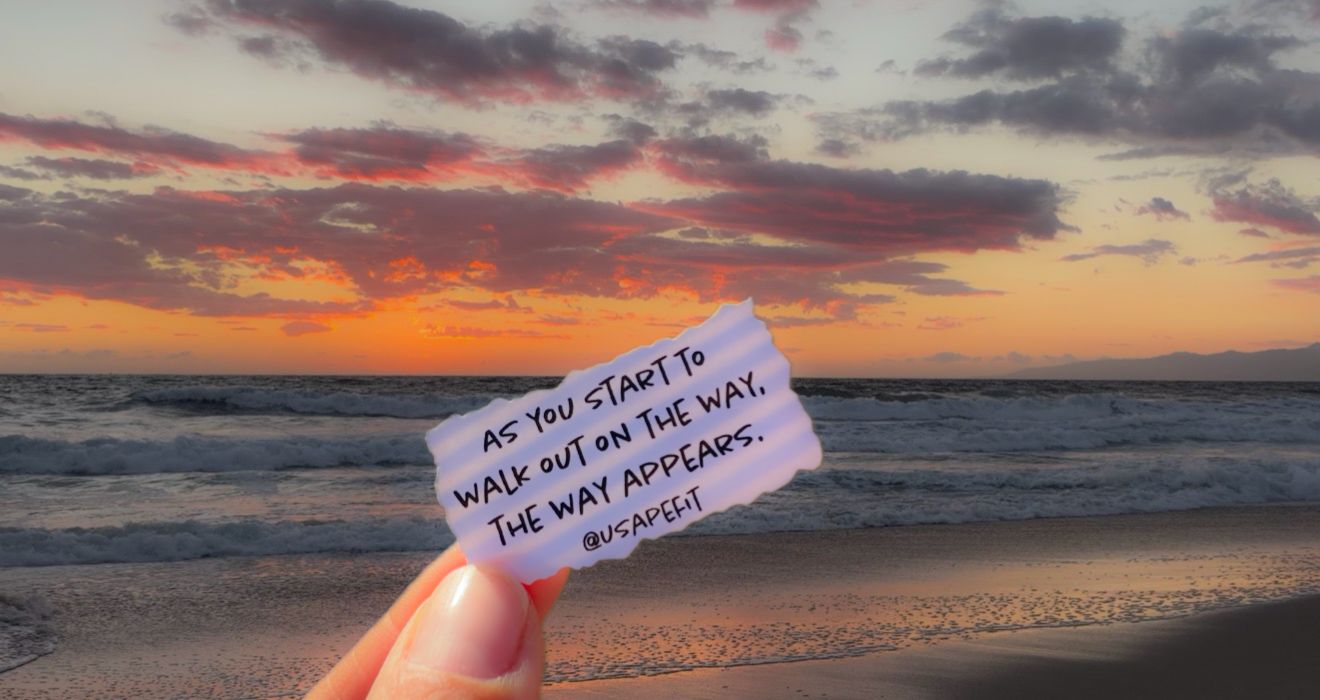 Hand holding a note reading 'As you start to walk out on the way, the way appears' against a vibrant sunset over the ocean.