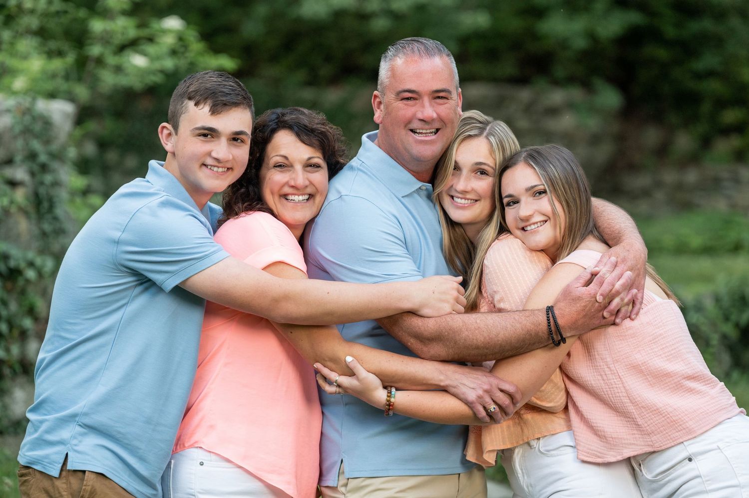 Happy family of six smiling and hugging outdoors, surrounded by lush green trees in a warm, candid portrait.