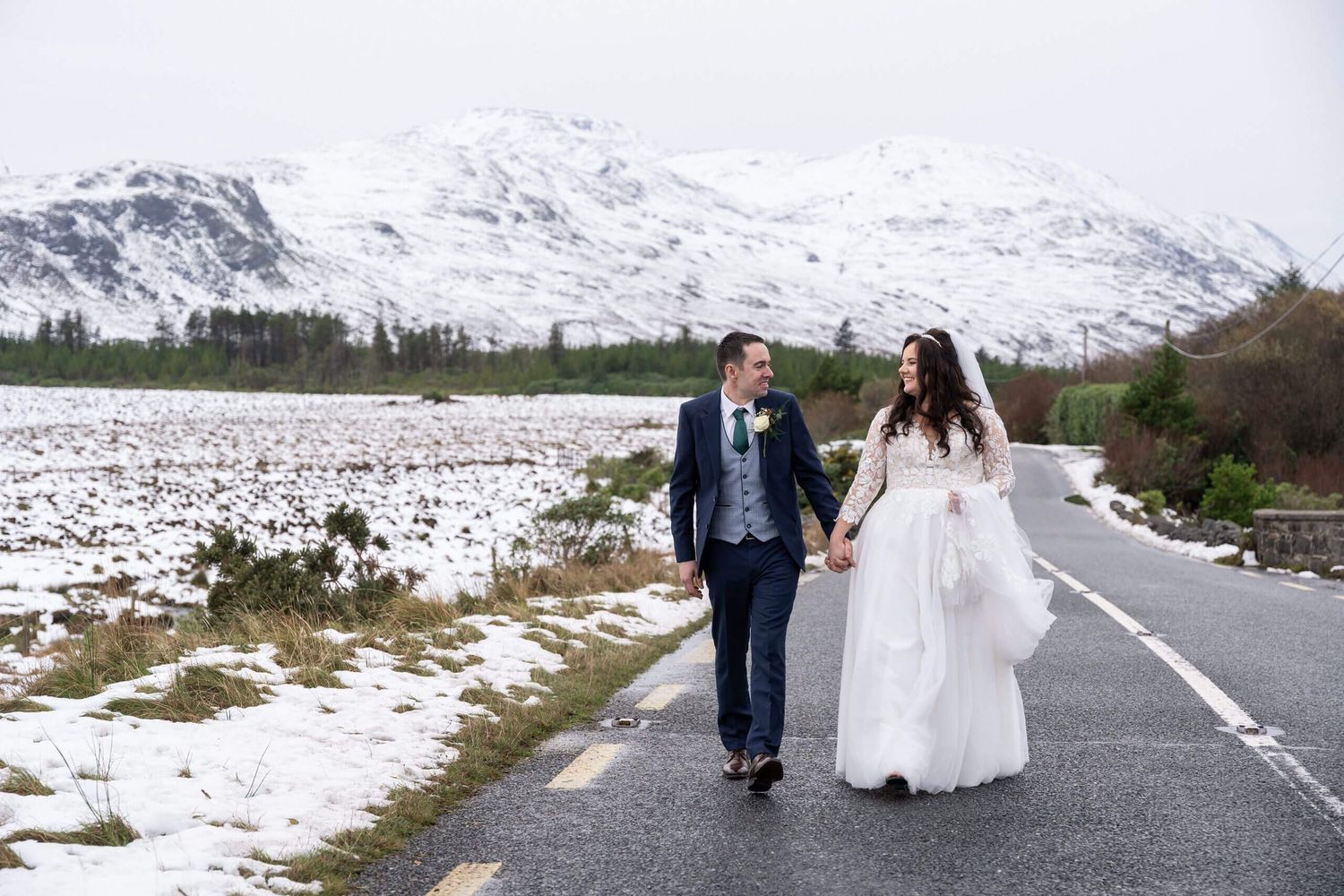 Bride and groom hold hands while walking down a quiet country road with snow-covered mountains and frosty fields behind them.