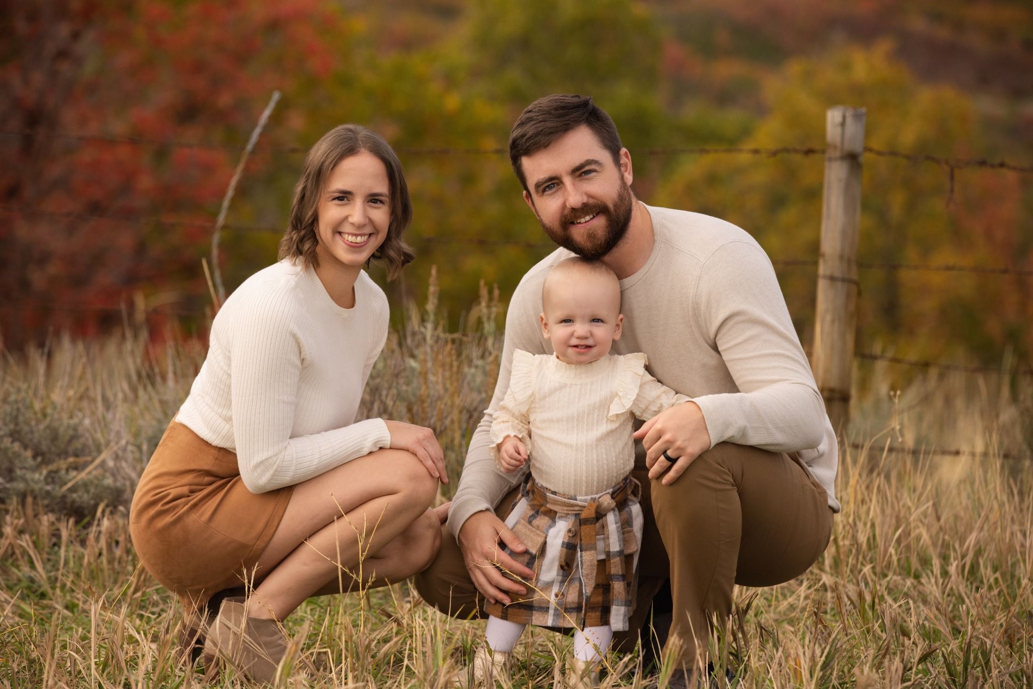 Parents crouching beside their young child in autumn grass with colorful foliage behind them
