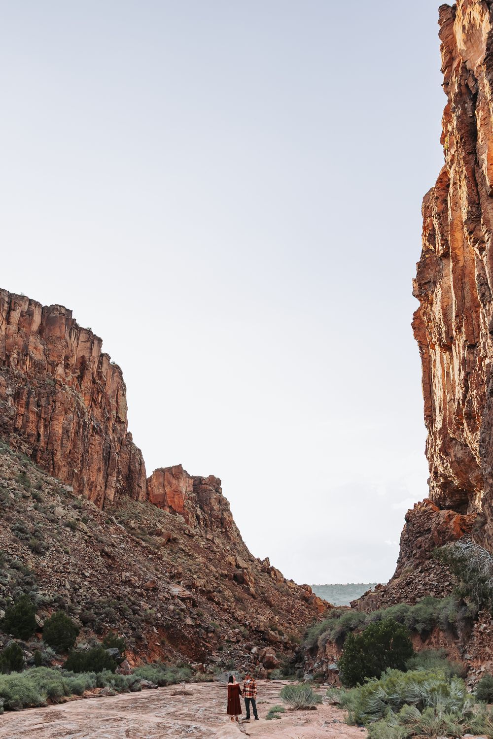 Two hikers stand small against towering red rock canyon walls in a vast desert landscape under a pale sky.