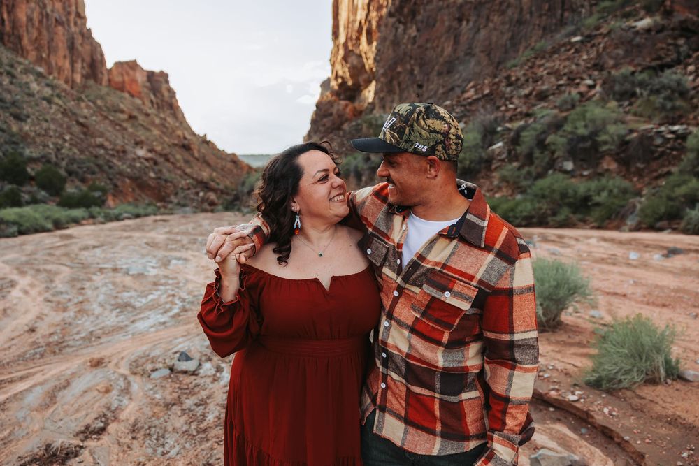 Couple smiling at each other in a red rock canyon, woman in red dress and man in plaid shirt and camo cap.