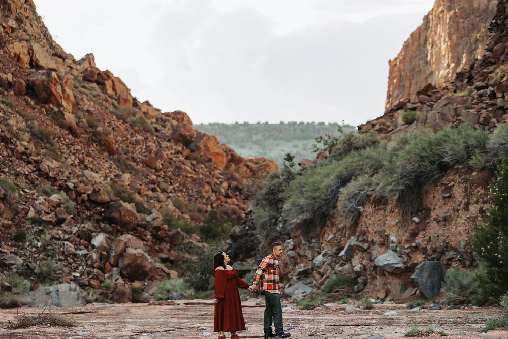 Couple holding hands in a scenic desert canyon with towering red rock walls and lush green shrubs.