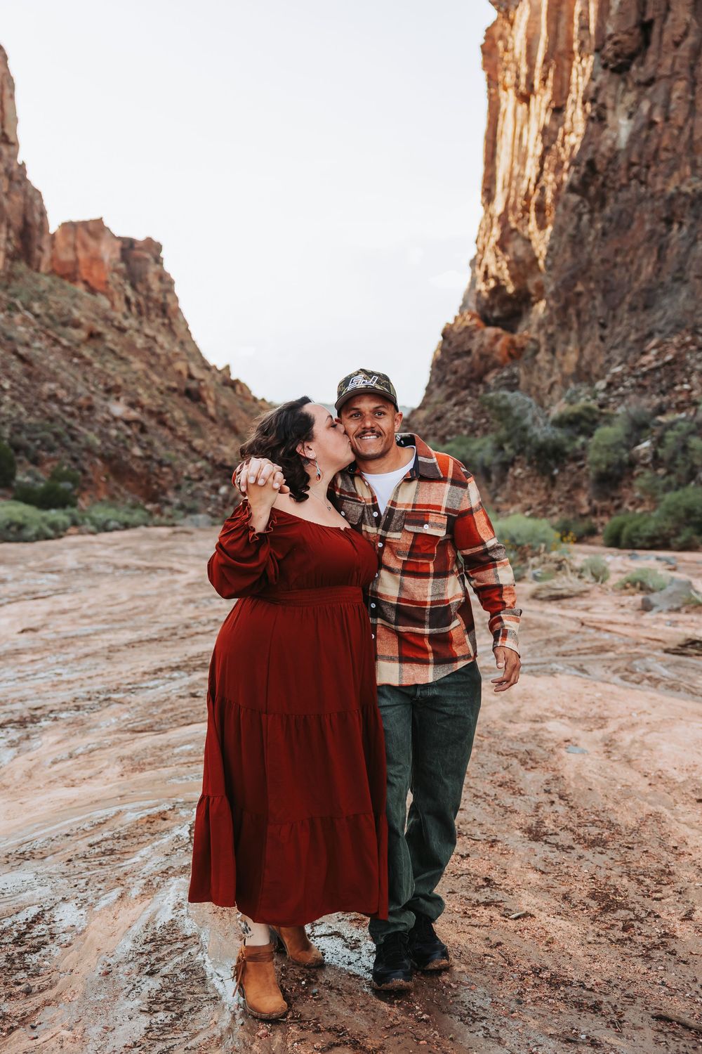 A couple shares a romantic kiss in a stunning red rock canyon, she wears a red dress and he wears a plaid shirt.