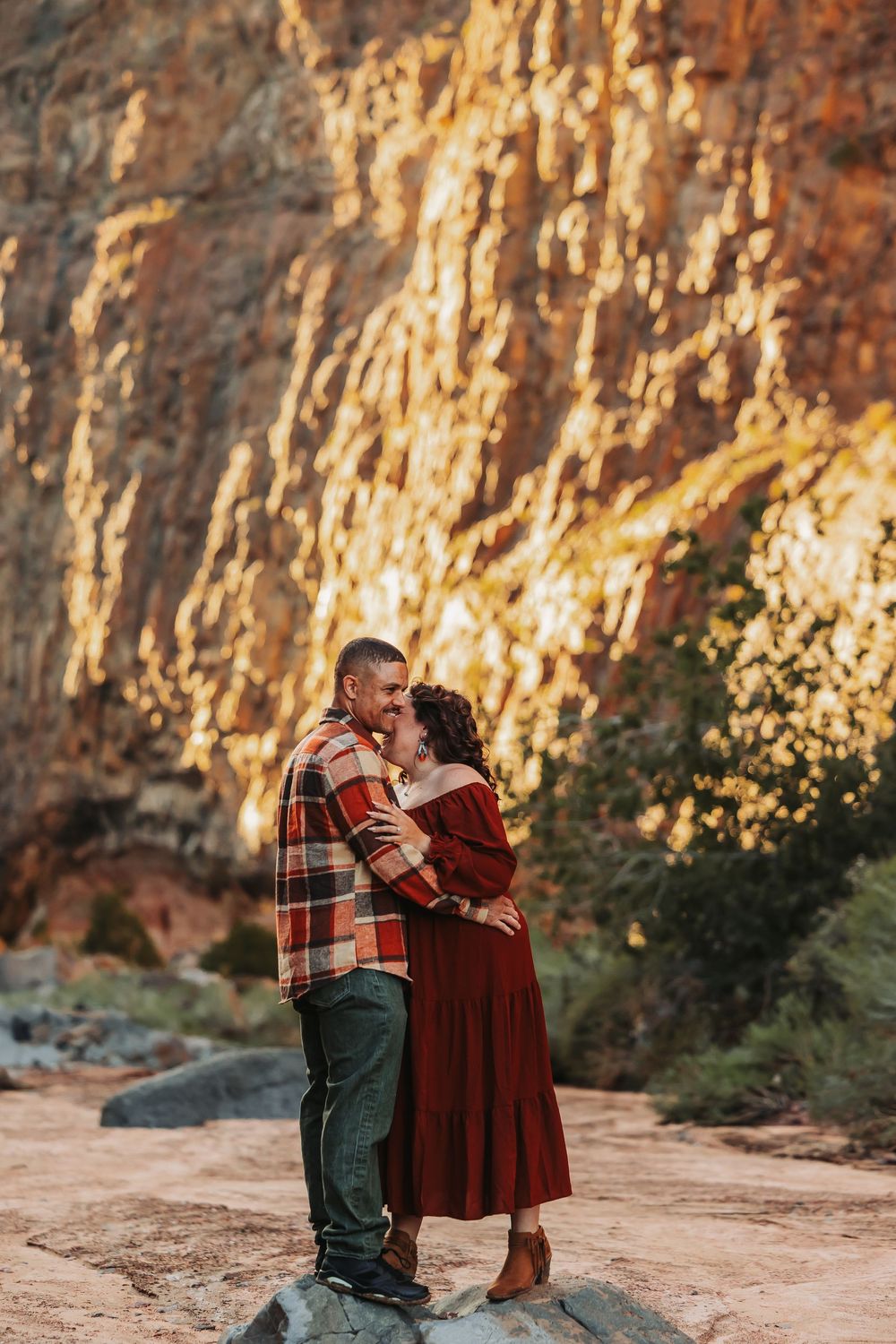 Couple embracing romantically in front of sunlit red rock canyon walls, woman in burgundy dress, man in plaid shirt.