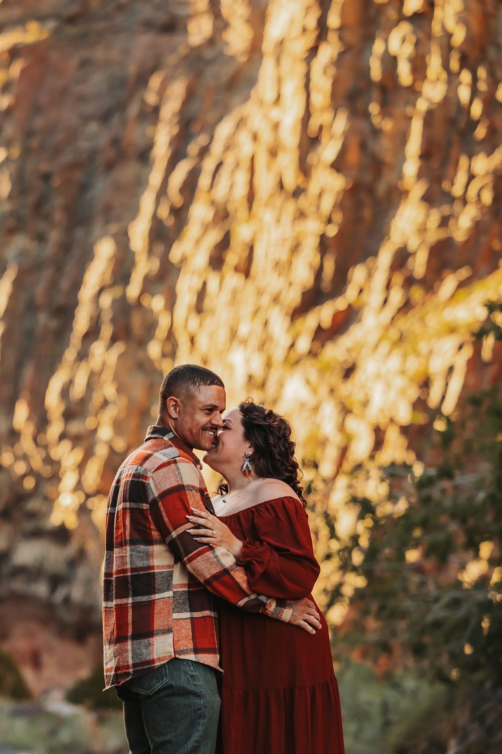Couple sharing a romantic kiss in front of golden sunlit red rock cliffs, wearing plaid and burgundy outfits.