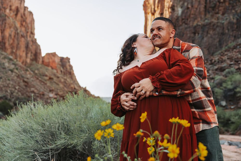 Couple embracing among yellow wildflowers with red rock canyon cliffs in background during outdoor engagement photo session.
