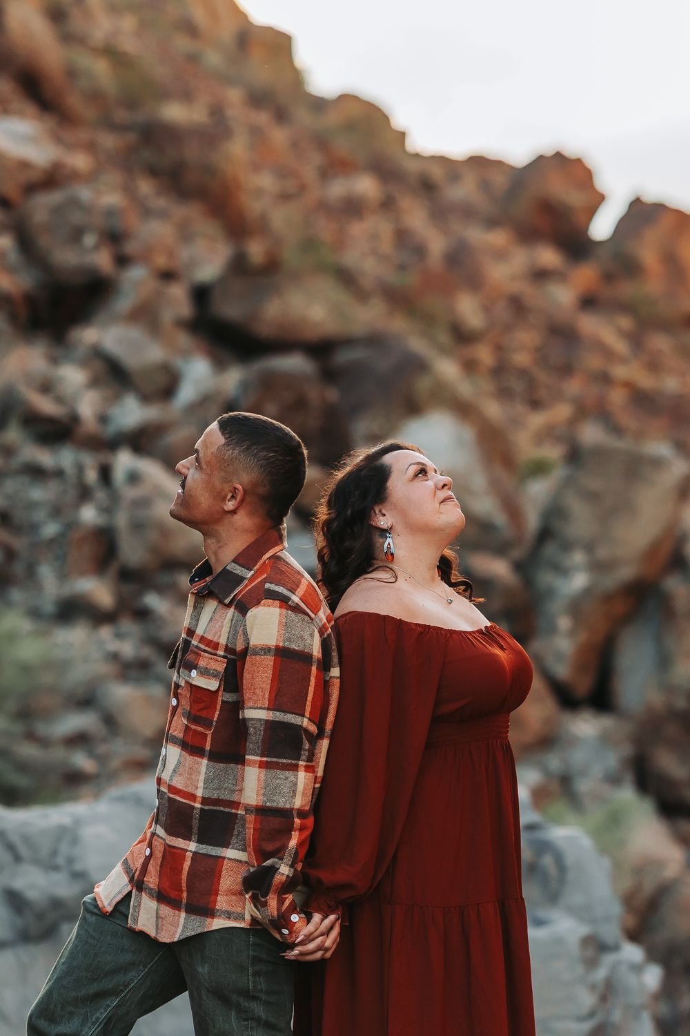 Couple standing back-to-back holding hands in front of rocky desert landscape, wearing plaid shirt and red off-shoulder dress.