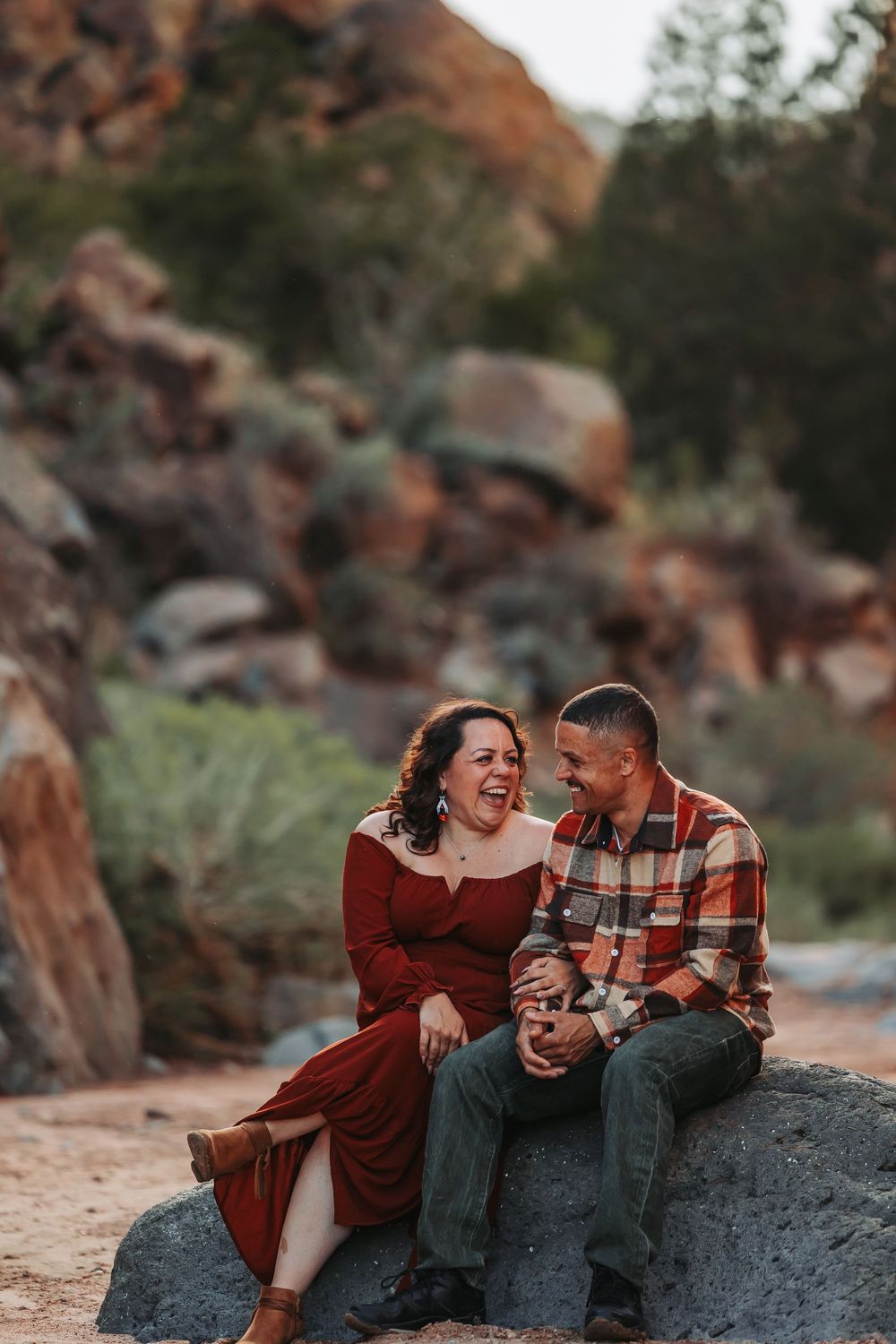 Couple laughing and holding hands while sitting on a rock with red canyon boulders in the background.