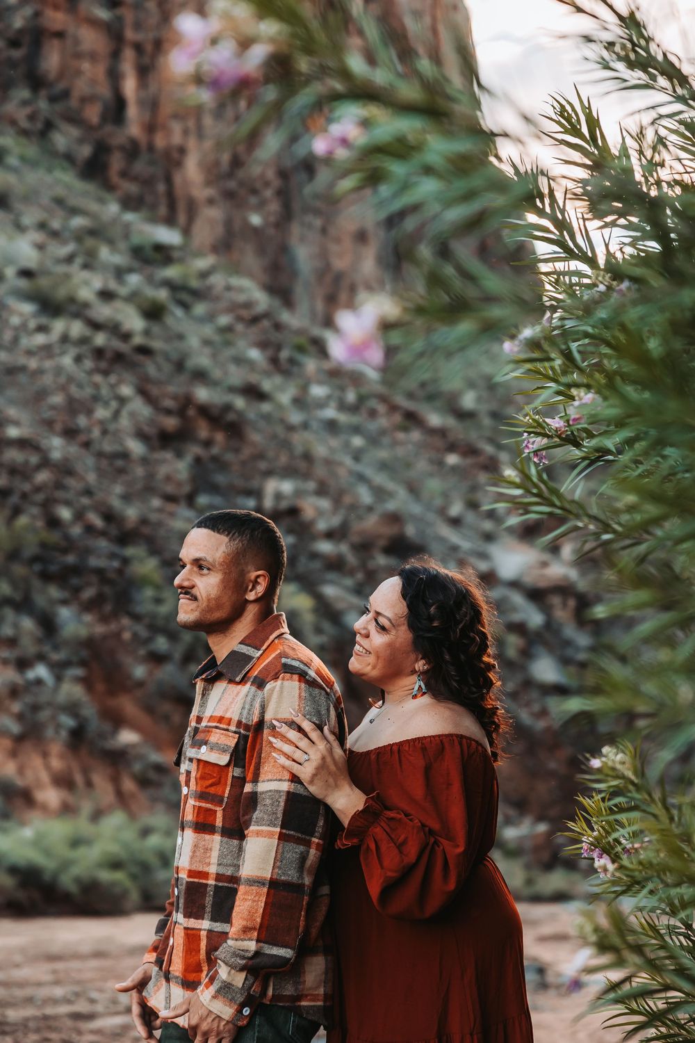 Couple embracing outdoors near lush greenery and rocky cliffs, woman in red dress resting hands on mans plaid shirt shoulders.
