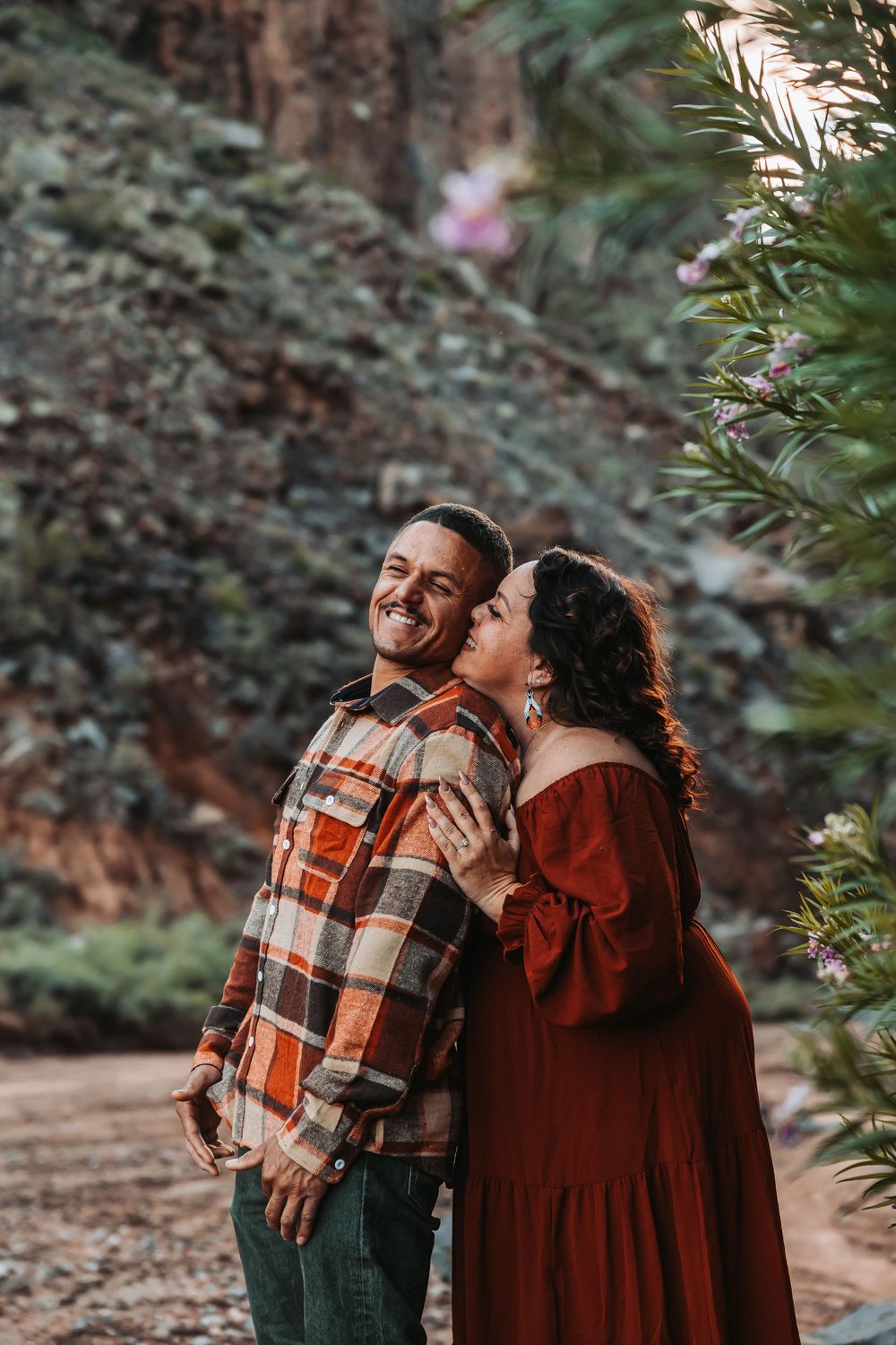 A couple shares a romantic moment outdoors, woman in red dress kissing mans cheek near flowering plants.