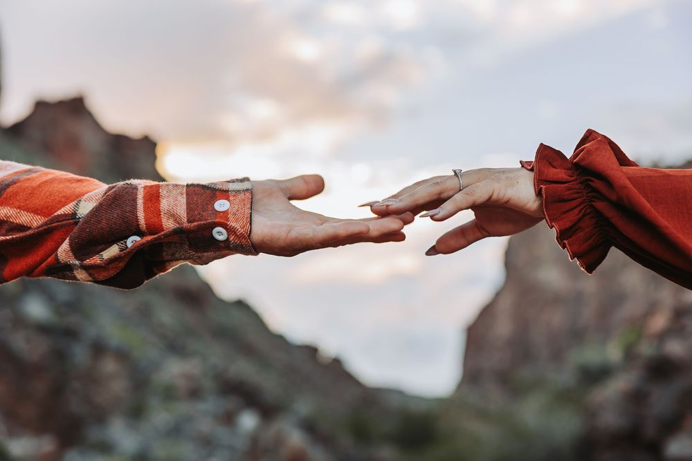 Two hands reaching toward each other against a scenic mountain backdrop at sunset, evoking connection and romance.