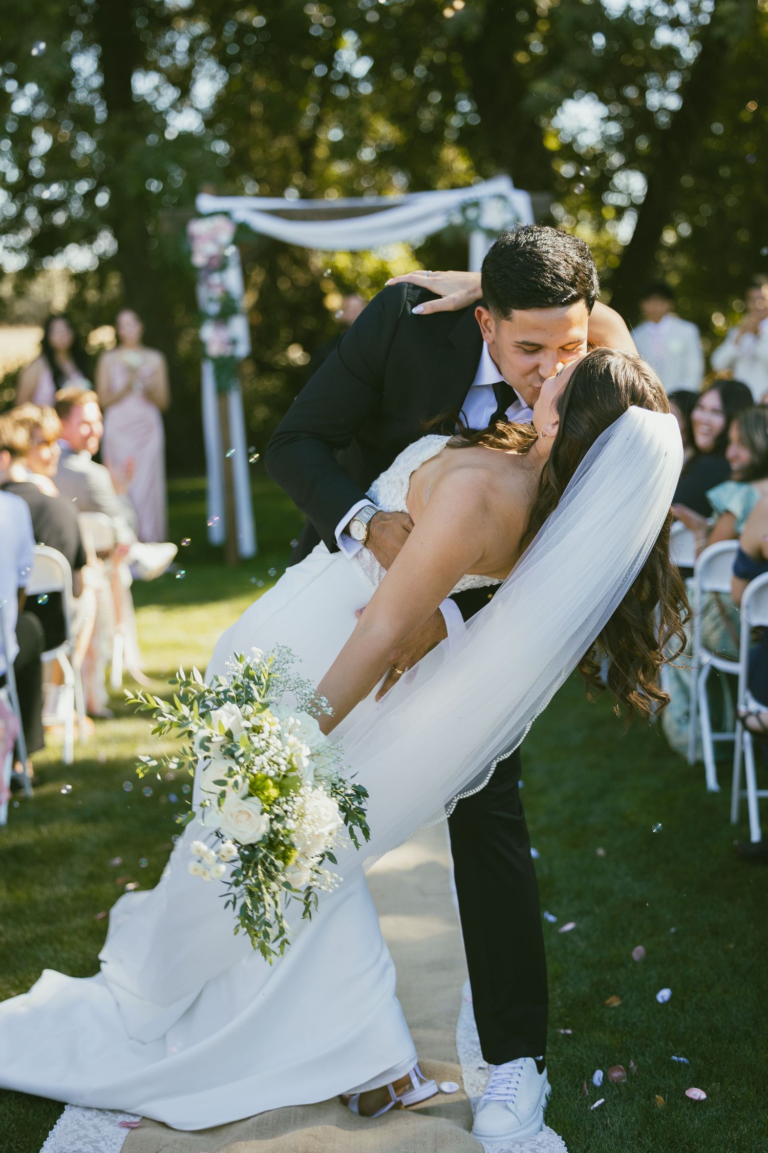 Groom dips bride for a kiss at outdoor wedding ceremony, bride holding white floral bouquet, guests seated on lawn.