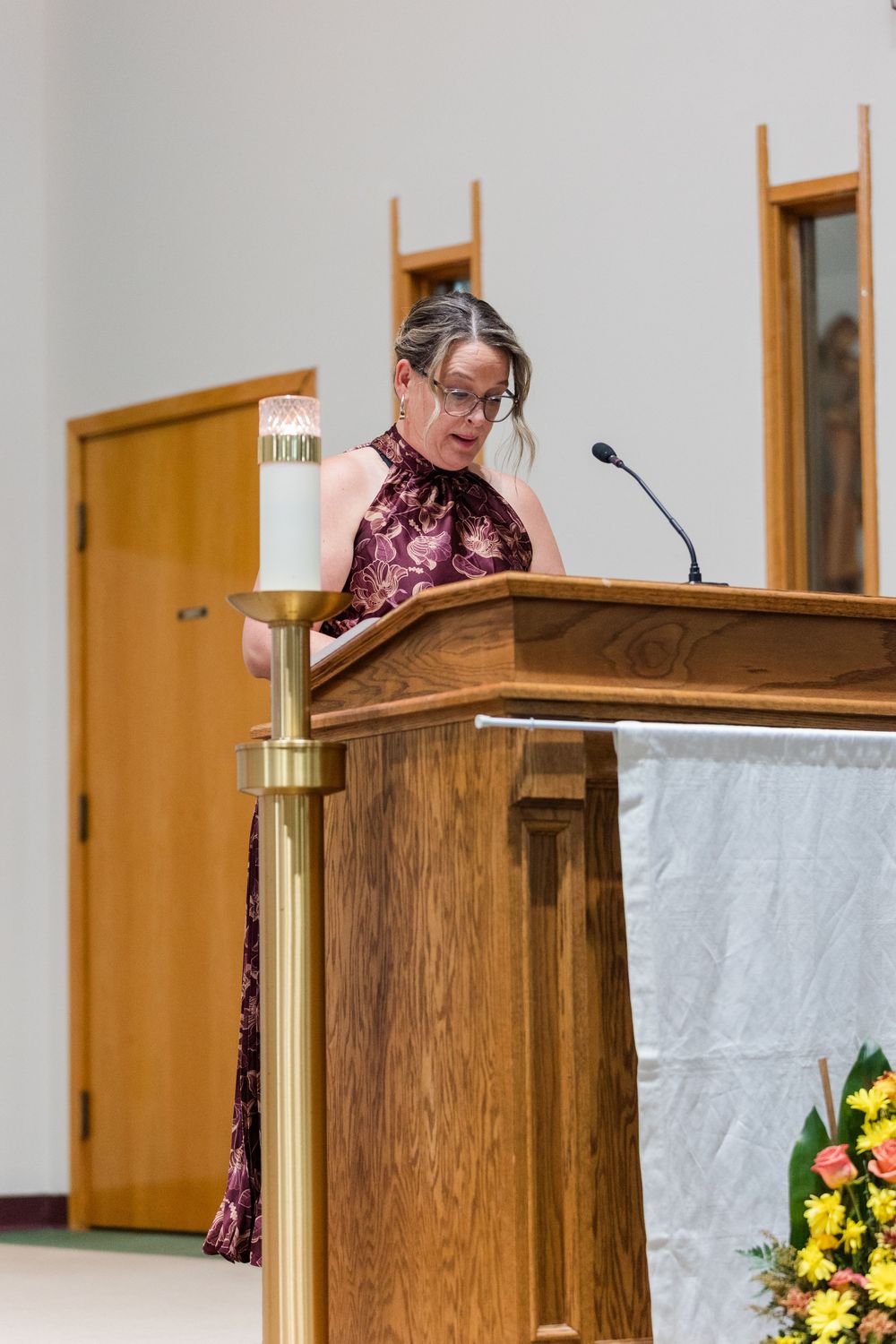 Woman in a burgundy patterned dress speaks into a microphone at a wooden church lectern beside a lit candle and yellow flowers.