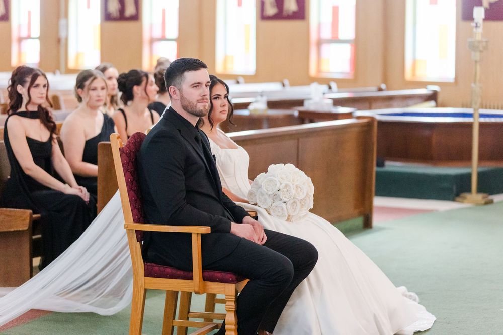 Bride in a white gown holding a bouquet sits beside the groom in a dark suit during a church wedding ceremony as bridesmaids look on.