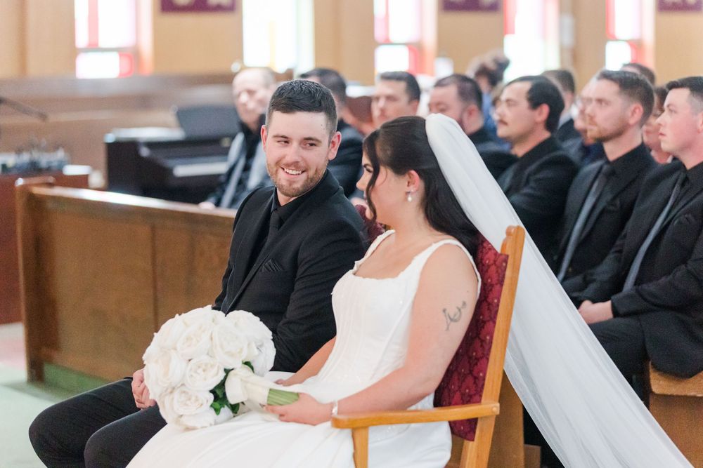 Smiling groom in a black suit sits beside a bride in a white gown holding a white rose bouquet during a church wedding ceremony with guests.