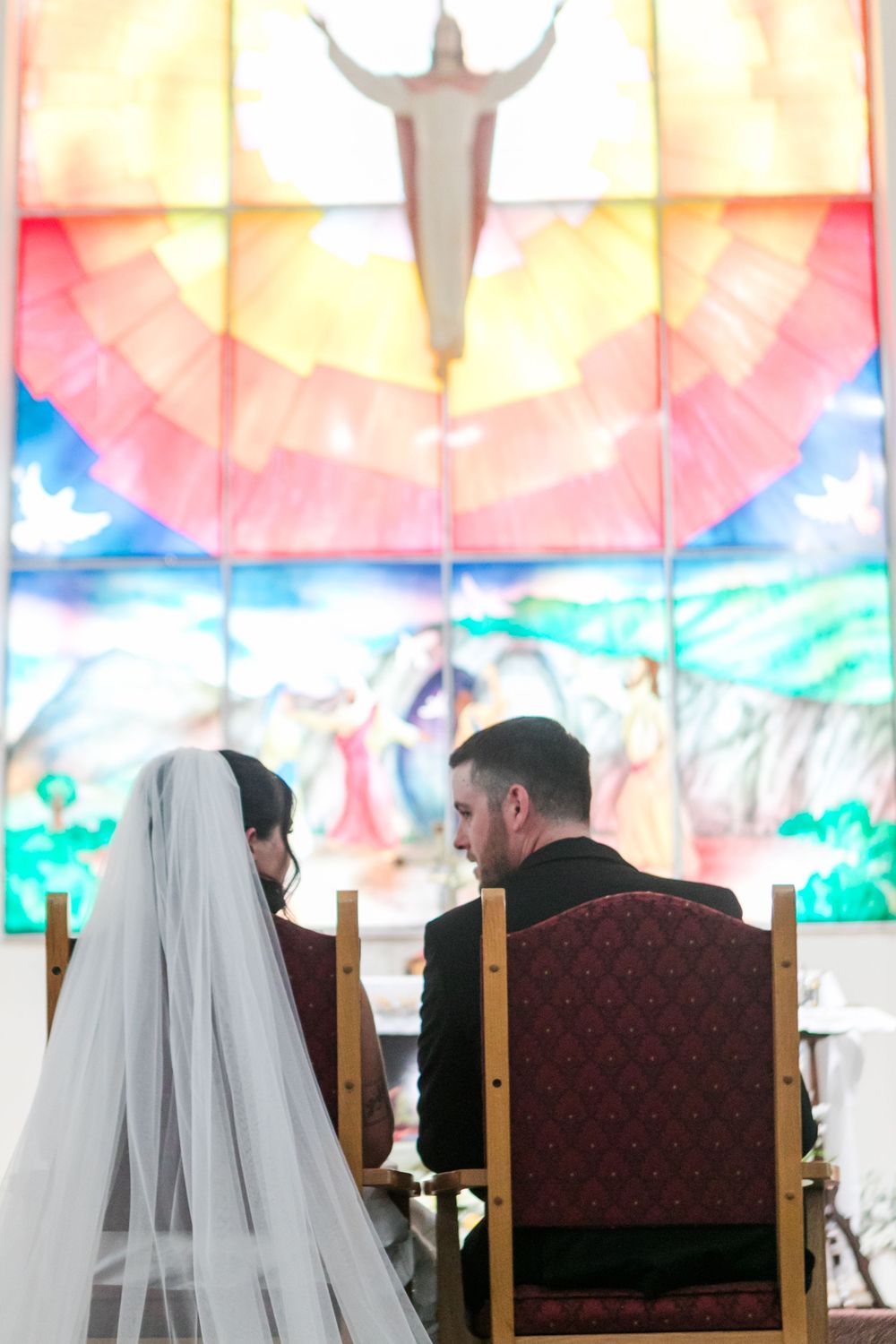 Bride in a long veil and groom in a dark suit sit before a colorful stained glass window with Jesus figure during a church wedding ceremony.