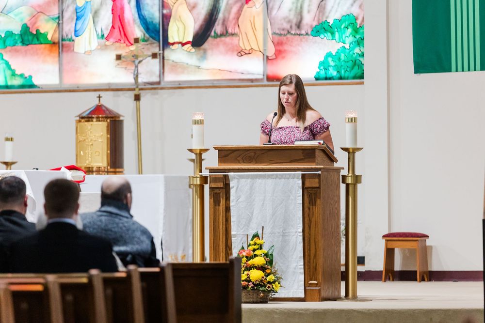 Young woman in a floral dress speaks at a wooden lectern beside candles and flowers during a church service, with attendees seated in front.