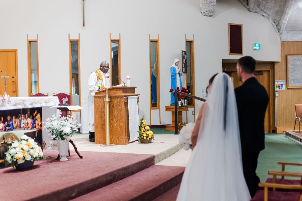 Bride in a white gown and veil stands beside a groom in a dark suit as a priest officiates their wedding ceremony at a church altar.