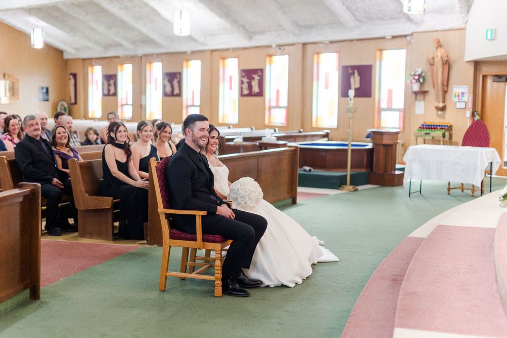 Bride in a white gown and groom in a dark suit sit in chairs facing the altar during a church wedding as guests watch from the pews.