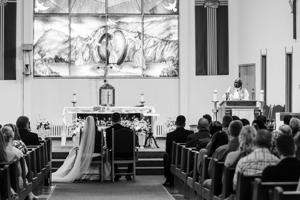 Black and white photo of a wedding ceremony in a church, with the bride and groom facing the altar as guests watch from the pews.