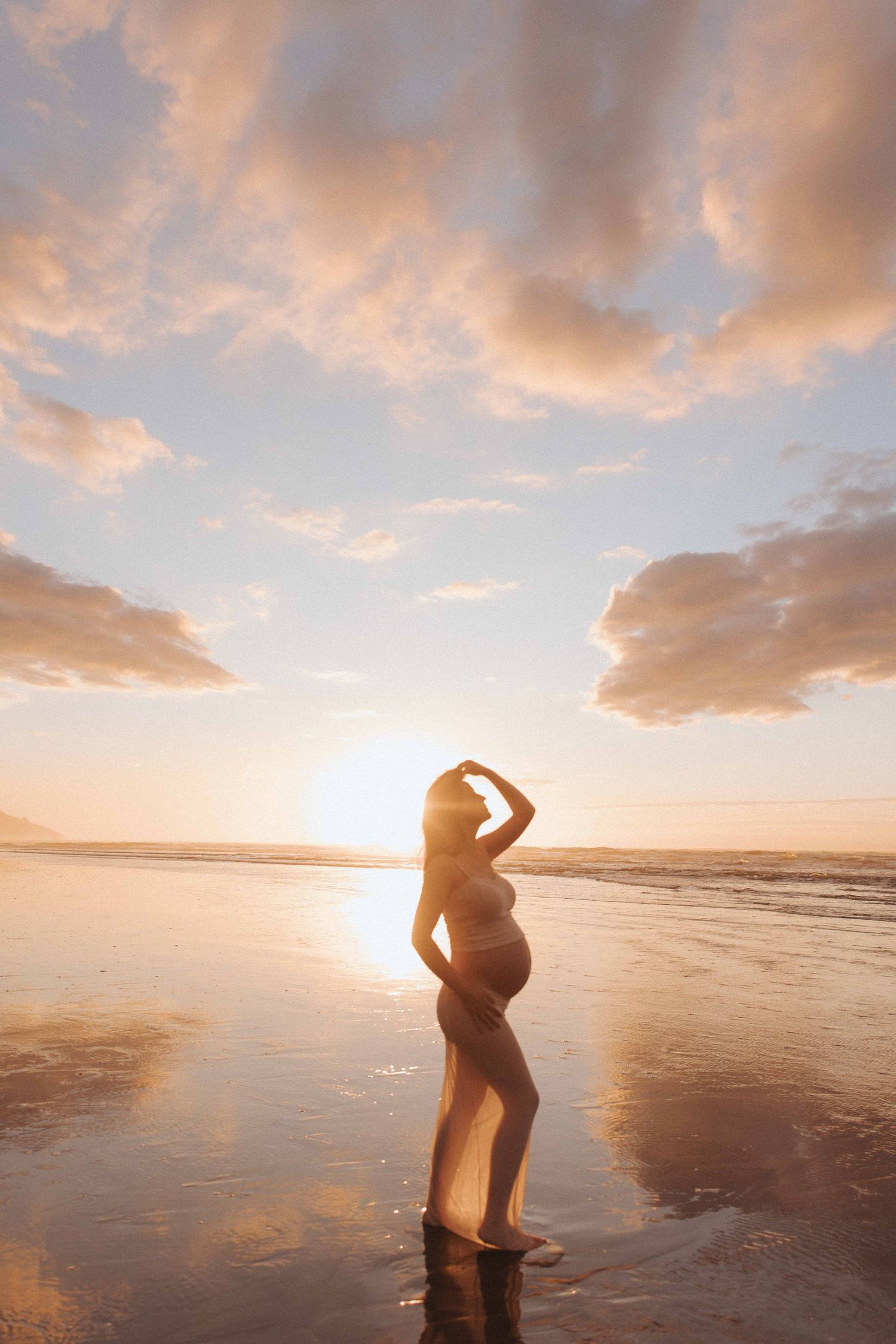 Pregnant woman in a sheer flowing gown poses on a wet beach at golden sunset, with glowing clouds reflected in the shallow water.