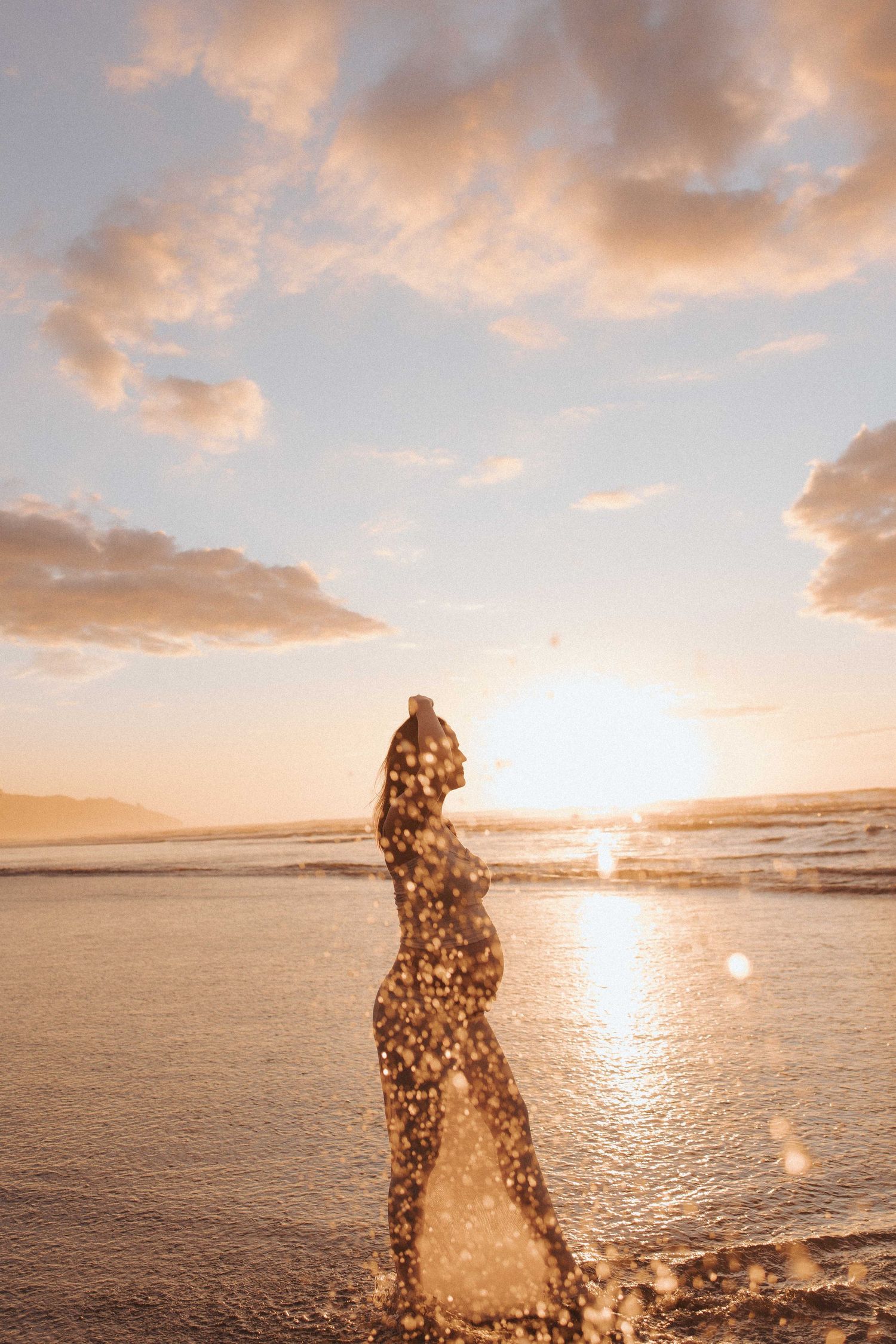 Silhouette of a pregnant woman standing in shallow ocean water at sunset, with golden light and sparkling water droplets under a cloudy sky.