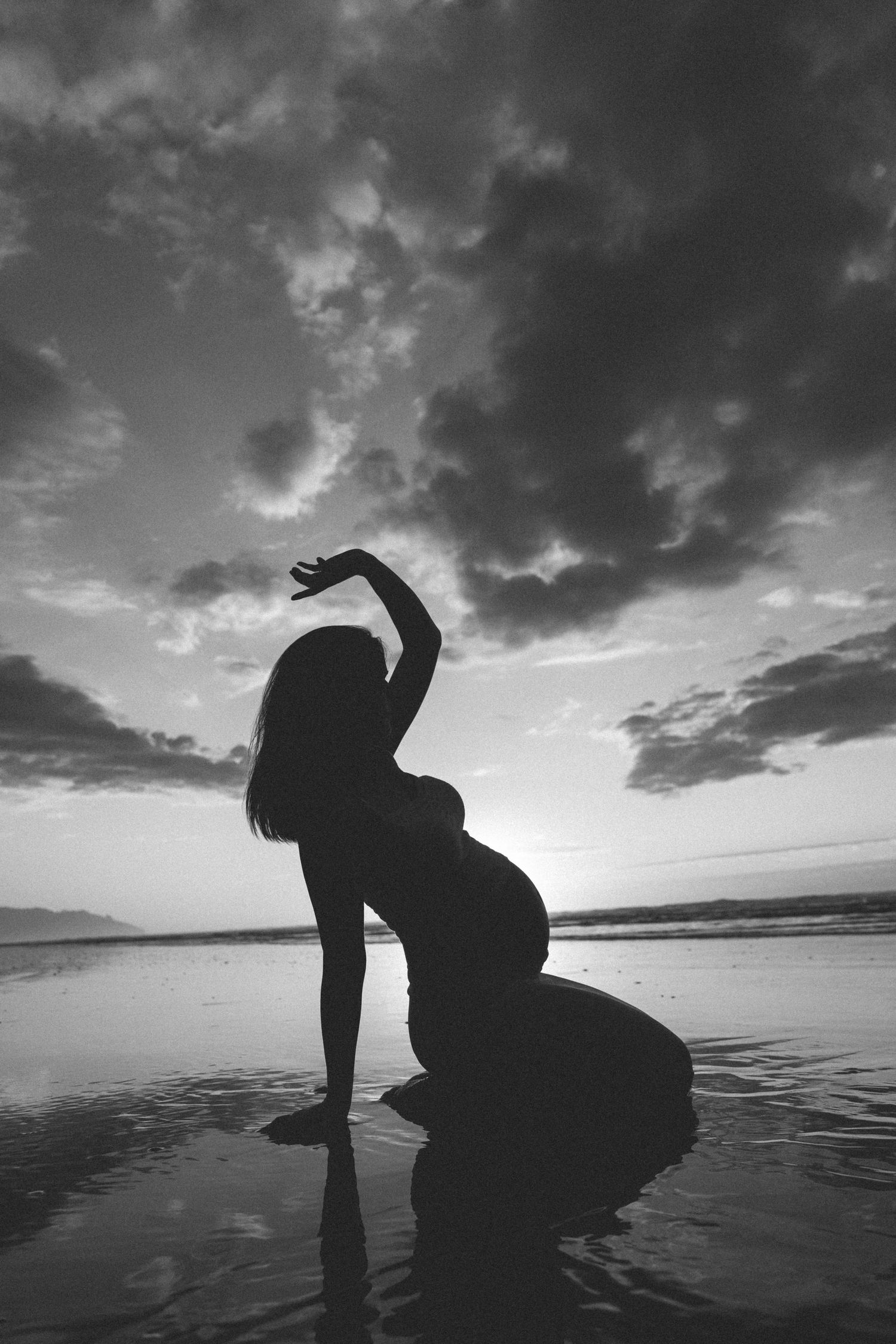 Black and white silhouette of a pregnant woman kneeling on a wet beach at sunset, one arm raised beneath dramatic clouds.