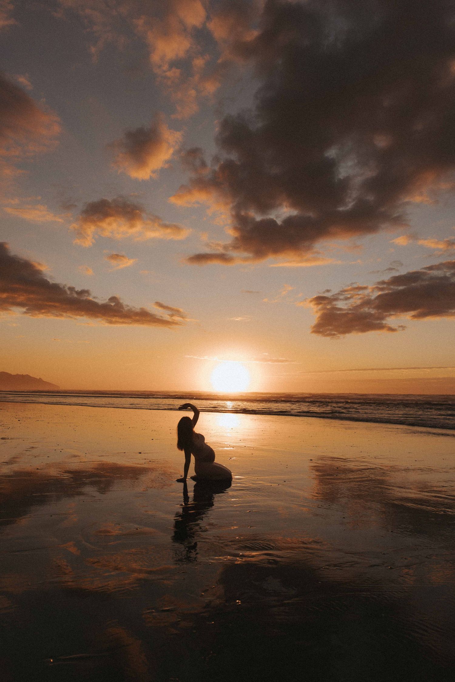 Silhouette of a pregnant woman kneeling on wet sand at sunset, with golden sun, dramatic clouds, and glowing reflections on the beach Raglan