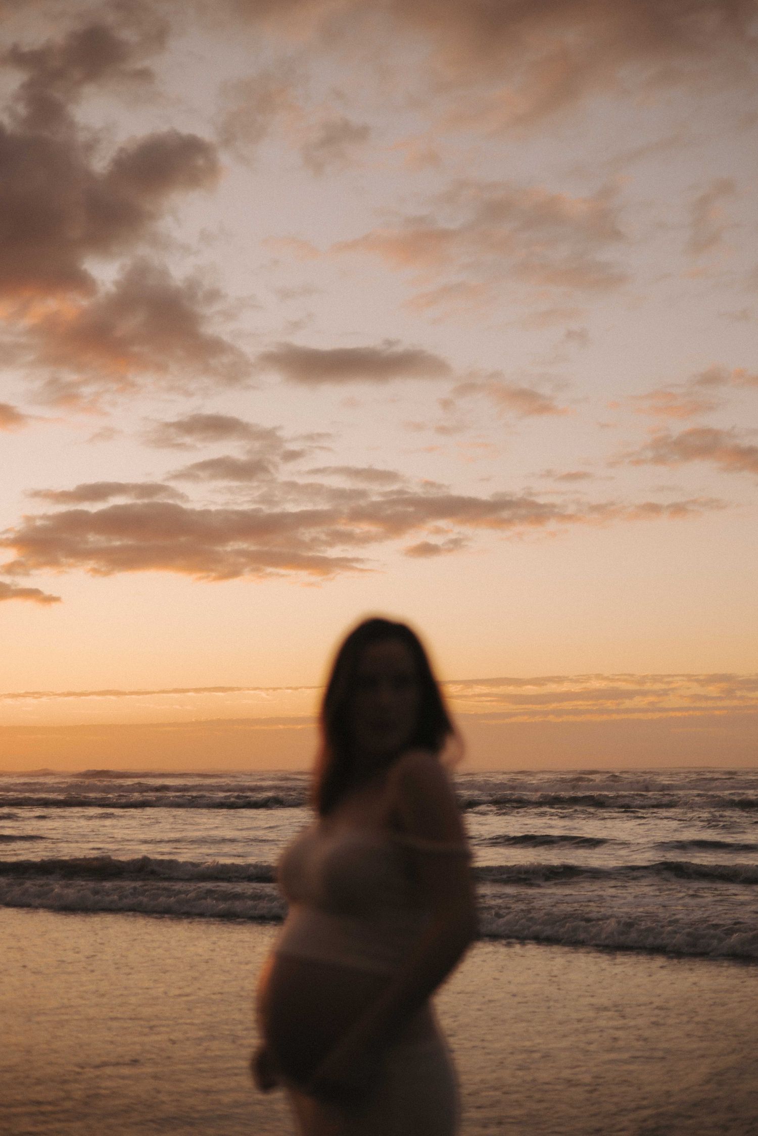 Blurred silhouette of a pregnant woman standing on a beach at sunset with soft orange clouds over the ocean.