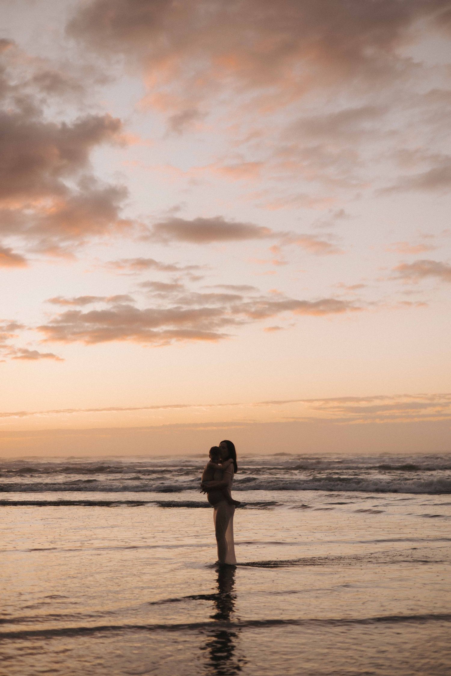 Silhouette of a pregnant woman holding a child while standing in shallow ocean water beneath a pink and orange sunset sky in Raglan