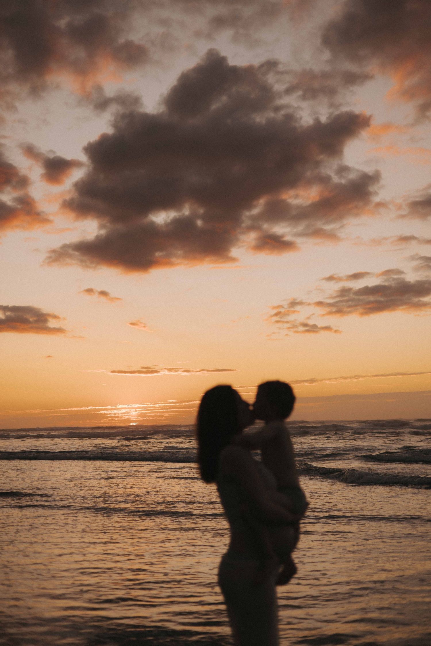 Silhouette of a mother holding and kissing her child on a beach at sunset, in Raglan New Zealand