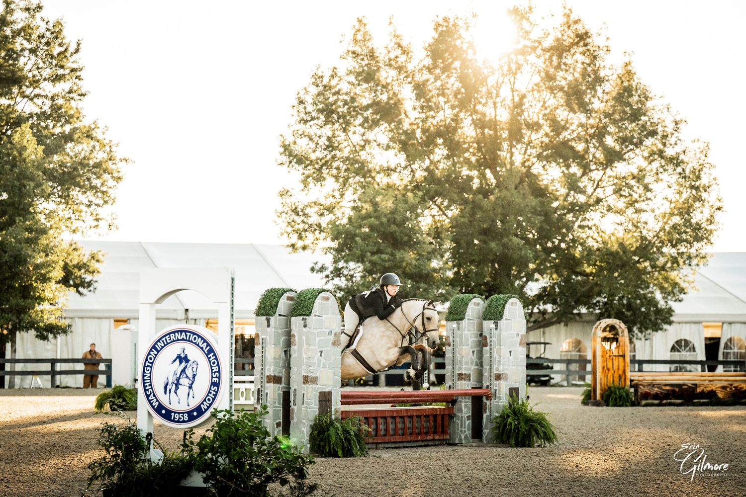 Equestrian rider in a helmet jumps a horse over a decorated obstacle at an outdoor show jumping event with trees and sunlight behind.
