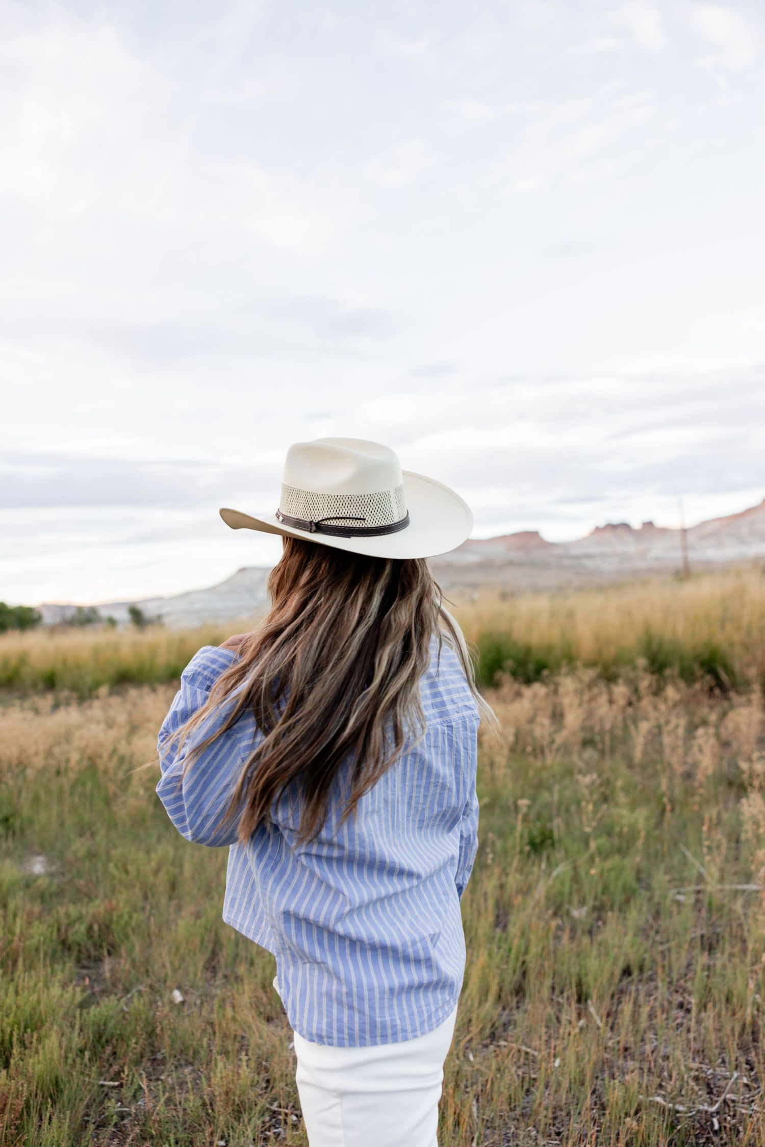 Woman seen from behind wearing a cream wide-brim hat and blue striped shirt, standing in a golden grass field with red rock hills beyond.