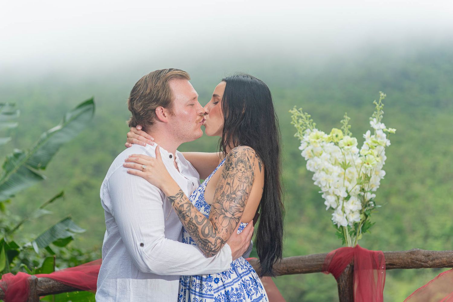 A couple shares a romantic kiss on a wooden balcony beside white flowers, with misty green mountains and lush foliage behind them.