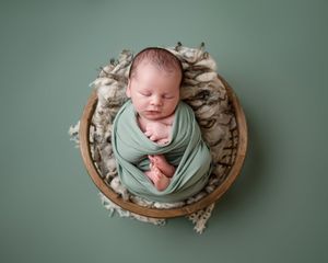 Sleeping newborn baby wrapped in a sage green swaddle resting in a round wooden bowl on a textured cream blanket against a green backdrop.