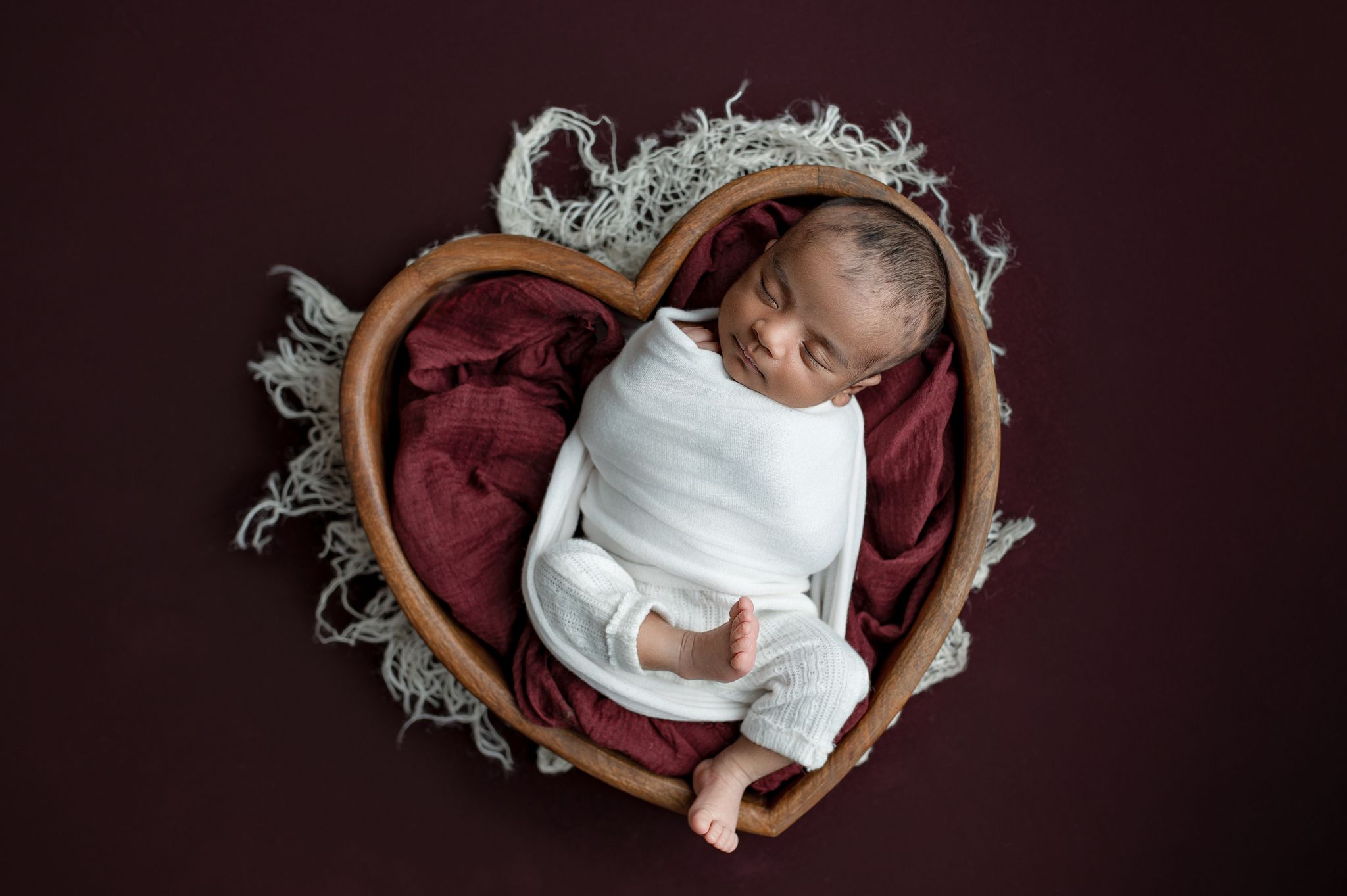 Newborn baby sleeping peacefully in a heart-shaped wooden bowl lined with a burgundy blanket and white wrap, photographed on a dark maroon b