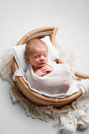 Sleeping newborn baby wrapped in white gauze lies in a crescent moon rattan bassinet on a cream knit blanket in a newborn photo session.