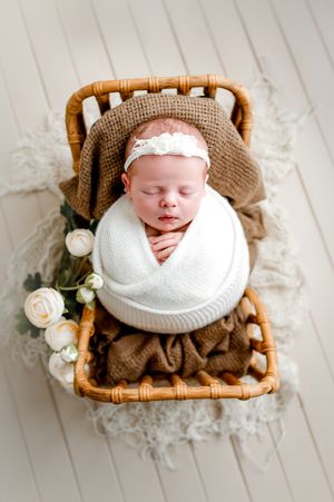 Newborn baby girl sleeping in a white swaddle with a lace headband inside a rattan bed, surrounded by cream ranunculus flowers.