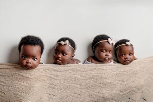 Four adorable baby girls with headbands peek over a cream knit blanket in a cute newborn photoshoot against a soft neutral backdrop.