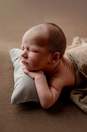 Sleeping newborn baby resting chin on folded hands atop a striped pillow, wrapped in a soft brown blanket during a newborn photo session.