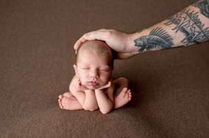 Sleeping newborn baby posed with chin resting on hands while a tattooed arm gently cradles the head on a brown backdrop.